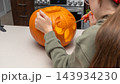 Child carving a pumpkin for Halloween with a small knife on a kitchen countertop, kitchen appliances visible in the background 143934230