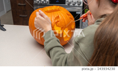 Child carving a pumpkin for Halloween with a small knife on a kitchen countertop, kitchen appliances visible in the background Child carving a pumpkin for Halloween with a small knife on a kitchen countertop, kitchen appliances visible in the background 143934230