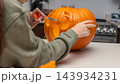 Person carving a pumpkin for Halloween in a kitchen, with a focus on the intricate design being created and kitchen appliances visible in the background 143934231