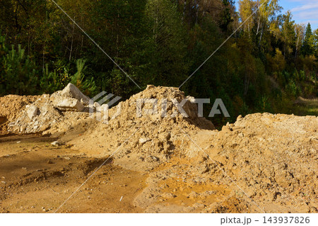 Huge soil heaps and heavy concrete blocks lie near edge of forest, creating rugged industrial dump site surrounded by green trees and autumn foliage 143937826