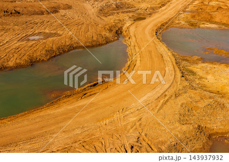 Dusty haul road winding through an open-pit sand quarry, featuring deep tire tracks and muddy water pools under natural sunlight, capturing industrial excavation site landscape aerial view 143937932