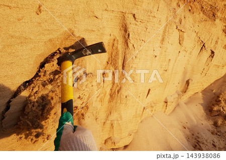 A gloved hand uses a small pickaxe to dig into a textured, golden sand embankment under warm sunlight, revealing layers and loose grains A gloved hand uses a small pickaxe to dig into a textured, golden sand embankment under warm sunlight, revealing layers and loose grains 143938086