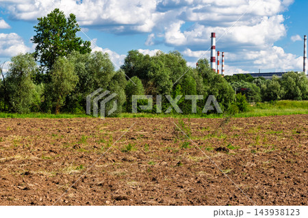 Freshly plowed dark soil field stretches before lush green forest trees under a bright blue sky with fluffy white clouds and industrial factory chimneys rising in the distance Freshly plowed dark soil field stretches before lush green forest trees under a bright blue sky with fluffy white clouds and industrial factory chimneys rising in the distance 143938123