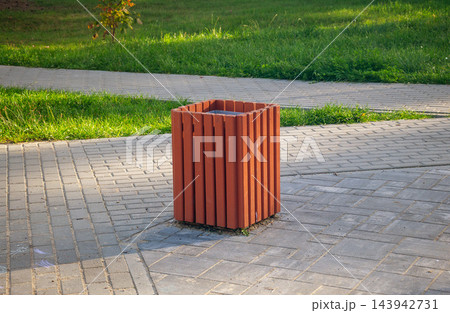 Modern wooden litter bin on stone tile pavement in public city park on autumn day Modern wooden litter bin on stone tile pavement in public city park on autumn day 143942731