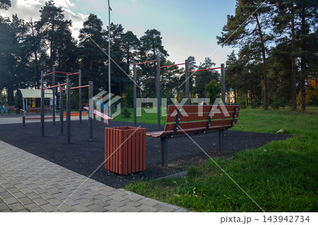 Empty wooden bench and pull up bars on modern calisthenics sports ground surrounded by green trees in public park outdoors 143942734