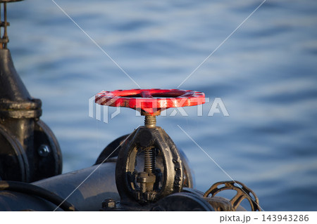 Red Metal Valve Wheel Mounted on Industrial Piping Against Blurred Water Background 143943286