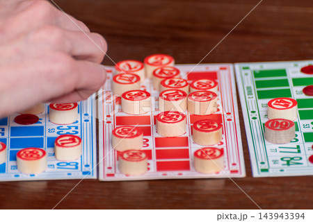 Player's hand marks a matching number on a bingo card with a wooden token during a casual indoor game, close up showing multiple cards and the thrill of winning Player's hand marks a matching number on a bingo card with a wooden token during a casual indoor game, close up showing multiple cards and the thrill of winning 143943394