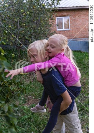 Two blond children, a boy giving a piggyback ride to a girl, are reaching for green apples hanging from a tree in a lush green garden with a brick house in the background Two blond children, a boy giving a piggyback ride to a girl, are reaching for green apples hanging from a tree in a lush green garden with a brick house in the background 143943530