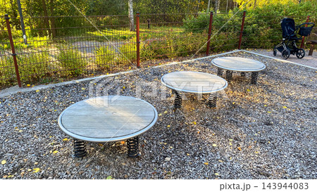 Three round spring-mounted balance platforms on outdoor playground with wood chip surface and baby stroller in background on sunny autumn day 143944083