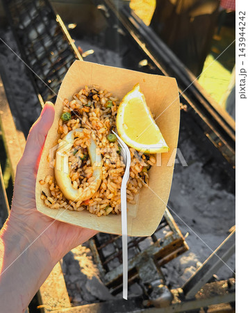 Portion of Seafood Paella with Squid and Lemon, takeaway food. Seafood paella with squid, green peas, lemon, disposable paper tray, held by hand. Spanish cuisine, street food, Outdoor Festival Portion of Seafood Paella with Squid and Lemon, takeaway food. Seafood paella with squid, green peas, lemon, disposable paper tray, held by hand. Spanish cuisine, street food, Outdoor Festival 143944242