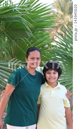Mother and son smile together outdoors, standing close beneath arching palm fronds. Relaxed family portrait reflects loving maternal bond, with both facing camera in bright daylight. Mother and son smile together outdoors, standing close beneath arching palm fronds. Relaxed family portrait reflects loving maternal bond, with both facing camera in bright daylight. 143945020