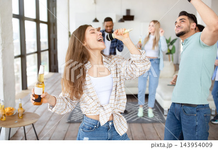 Young blonde lady partying with her diverse friends, singing karaoke into mic, dancing and drinking beer at home. Happy millennials socializing with alcohol drinks, moving to music, having fun indoors Young blonde lady partying with her diverse friends, singing karaoke into mic, dancing and drinking beer at home. Happy millennials socializing with alcohol drinks, moving to music, having fun indoors 143954009