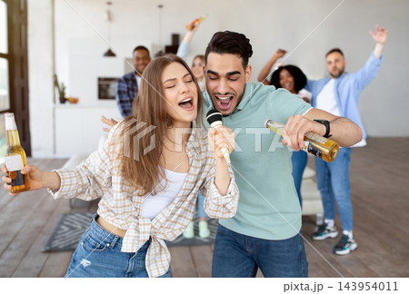 Young multiracial couple singing song together into microphone, drinking beer, partying with their friends at home. Happy Latin guy and Caucasian woman performing karaoke on student party Young multiracial couple singing song together into microphone, drinking beer, partying with their friends at home. Happy Latin guy and Caucasian woman performing karaoke on student party 143954011