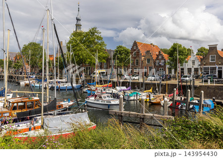 Horizontal perspective of town hall clock tower harbor marina sailboats traditional Dutch brick houses wooden posts blue sky Veere Zeeland Netherlands European travel commercial 143954430