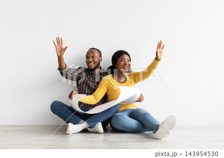 Happy Black Couple Sitting On Floor In New Flat, Holding Blueprints And Discussing Interior Design, Excited Young African American Spouses With Building Plan Imagining Style Of Their Apartment Happy Black Couple Sitting On Floor In New Flat, Holding Blueprints And Discussing Interior Design, Excited Young African American Spouses With Building Plan Imagining Style Of Their Apartment 143954530