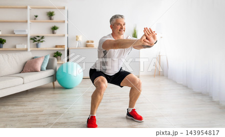Strength workout concept. Active mature man doing squats at home, exercising in living room, panorama, free space. Athletic man in sports clothes having domestic training during covid-19 lockdown Strength workout concept. Active mature man doing squats at home, exercising in living room, panorama, free space. Athletic man in sports clothes having domestic training during covid-19 lockdown 143954817