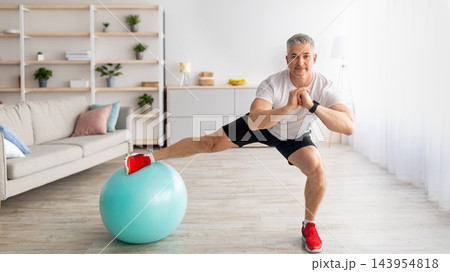 Happy sporty mature man doing side lunges, using fitness ball, working out, training in the morning during quarantine at home in living room interior, panorama with copy space 143954818