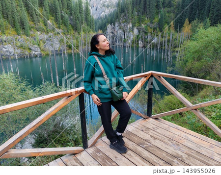 A traveler in a teal jacket pauses at a lakeside viewpoint, enjoying a still landscape of blue-green water, bare trunks and conifer-covered hills 143955026