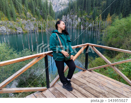 A woman rests against a wooden viewing-platform rail above a turquoise lake filled with bare tree trunks, framed by steep forested slopes A woman rests against a wooden viewing-platform rail above a turquoise lake filled with bare tree trunks, framed by steep forested slopes 143955027