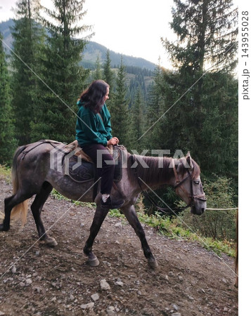 A gray horse carries a woman past tall evergreen trees, showing the steady pace of a recreational ride through a mountain landscape A gray horse carries a woman past tall evergreen trees, showing the steady pace of a recreational ride through a mountain landscape 143955028