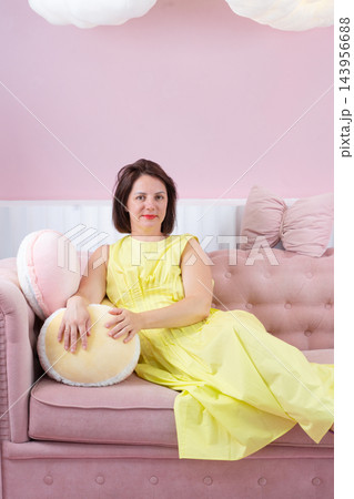 Young woman reclining on pink sofa in pastel living room wearing yellow dress and purple sandals, eyes closed in calm fashion pose 143956688