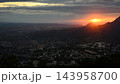 Wide landscape view of a city of Pyatigorsk during sunset. Bright orange sun setting behind mountain peaks with dark silhouettes. Dramatic cloudy sky over urban town 143958700