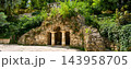 Man-made stone grotto with classical columns in a lush green park in Pyatigorsk.. Artificial cave entrance with pillars and overgrown foliage. Historical garden landscape architecture in summer 143958705