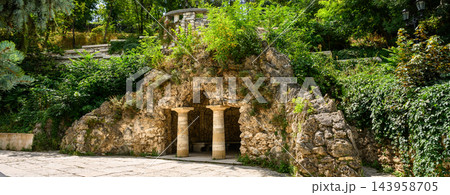 Man-made stone grotto with classical columns in a lush green park in Pyatigorsk.. Artificial cave entrance with pillars and overgrown foliage. Historical garden landscape architecture in summer 143958705