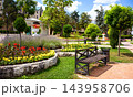 Landscaped public park with a wooden bench and colorful flower beds. Urban garden featuring topiary trees, stone paths, and green lawns on a sunny summer day in Pyatigorsk, Russia 143958706