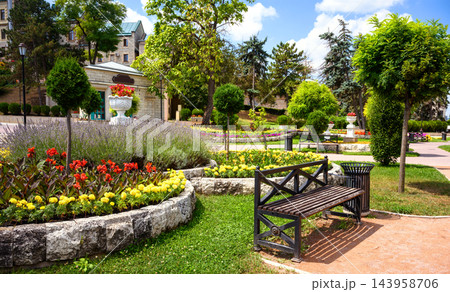Landscaped public park with a wooden bench and colorful flower beds. Urban garden featuring topiary trees, stone paths, and green lawns on a sunny summer day in Pyatigorsk, Russia Landscaped public park with a wooden bench and colorful flower beds. Urban garden featuring topiary trees, stone paths, and green lawns on a sunny summer day in Pyatigorsk, Russia 143958706