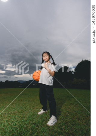 Young girl standing on green field holding orange basketball with cloudy sky and trees in background confidently posing Young girl standing on green field holding orange basketball with cloudy sky and trees in background confidently posing 143970039