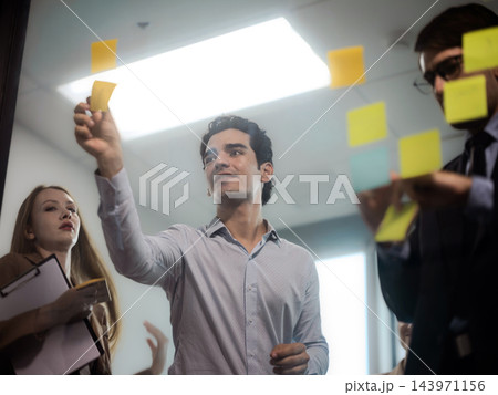 Creative business team brainstorming and organizing ideas with colorful sticky notes on a glass board during an office meeting. 143971156