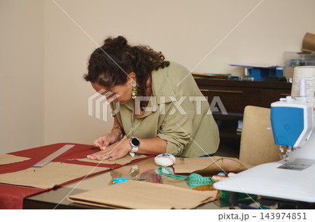 Focused woman pins a sewing pattern onto red fabric while creating handmade fashion designs at a home tailoring workshop table Focused woman pins a sewing pattern onto red fabric while creating handmade fashion designs at a home tailoring workshop table 143974851