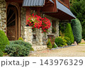 Rustic stone cottage entrance framed by hanging red flowers and carefully trimmed garden shrubs 143976329