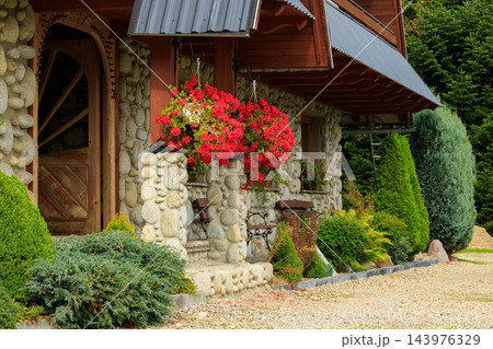 Rustic stone cottage entrance framed by hanging red flowers and carefully trimmed garden shrubs 143976329