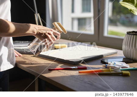 Person sprays water onto textured canvas surrounded by painting supplies Person sprays water onto textured canvas surrounded by painting supplies 143977646