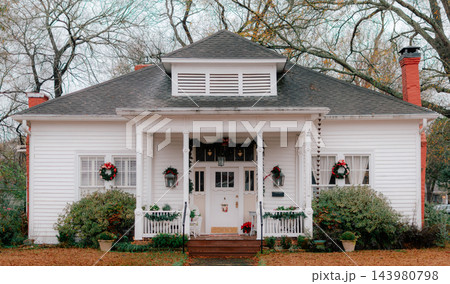 JEFFERSON, TEXAS: A historic white wooden cottage with a front porch, dormer window, and red brick chimney decorated with Christmas wreaths and festive greenery. JEFFERSON, TEXAS: A historic white wooden cottage with a front porch, dormer window, and red brick chimney decorated with Christmas wreaths and festive greenery. 143980798