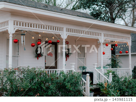 JEFFERSON, TEXAS: A white historic home's front porch decorated for the holidays with large red hanging ornament balls, string lights, and festive holiday decor. 143981115