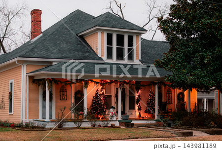 JEFFERSON, TEXAS: A warm light-orange historic house decorated for Christmas at dusk, featuring illuminated string lights, festive garland, and Christmas trees on the porch. JEFFERSON, TEXAS: A warm light-orange historic house decorated for Christmas at dusk, featuring illuminated string lights, festive garland, and Christmas trees on the porch. 143981189