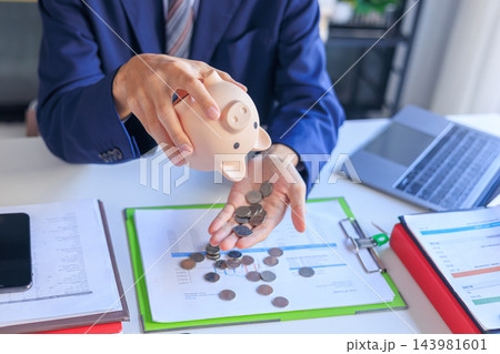 Businessman Shaking Coins Out of a Piggy Bank Onto Table with Financial Documents. Financial Planning, Personal Capital Allocation, and Wealth Investment Concept. 143981601