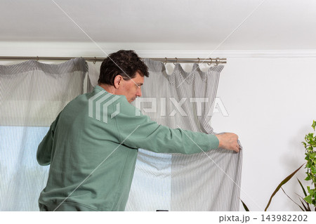 A man is hanging a curtain on the rod after washing it. It is spring, and he is doing household chores in a room with a window and some greenery A man is hanging a curtain on the rod after washing it. It is spring, and he is doing household chores in a room with a window and some greenery 143982202