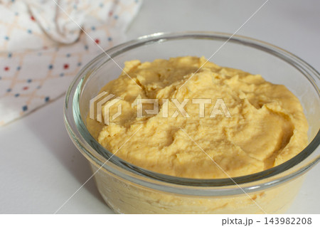 Hummus fills a glass bowl on a kitchen counter, with a patterned cloth nearby as someone prepares food at home during the day 143982208