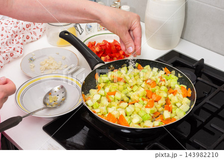 A hand adds seasoning to diced squash and other vegetables in a pan on a kitchen stove during the preparation of squash caviar 143982210