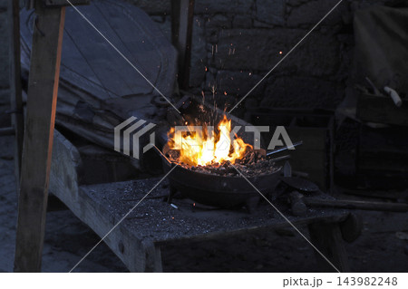 Medieval forge with bright flames and large leather bellows behind the hearth, surrounded by charcoal and traditional blacksmithing equipment in a rustic smithy at a historical fair. Medieval forge with bright flames and large leather bellows behind the hearth, surrounded by charcoal and traditional blacksmithing equipment in a rustic smithy at a historical fair. 143982248