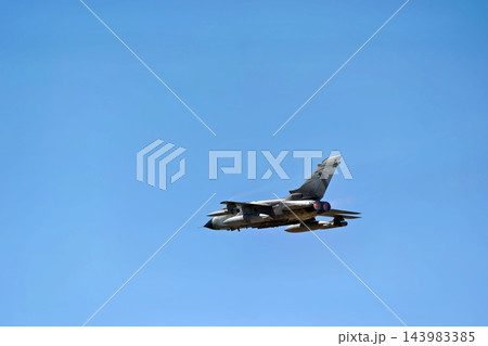 Military combat jet flies across a bright blue sky with glowing engine exhausts and mounted fuel tanks during an aerial flight demonstration Military combat jet flies across a bright blue sky with glowing engine exhausts and mounted fuel tanks during an aerial flight demonstration 143983385