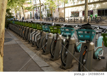 Row of public rental bicycles with green and teal baskets parked along city sidewalk beneath leafy trees and sunlit shadows, street view with railing and storefront background 143986544