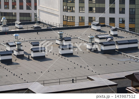 Rooftop view of flat roof features vents skylights and metal chimneys against modern beige and gray building facade under clear daylight with metal railings and concrete parapets 143987512