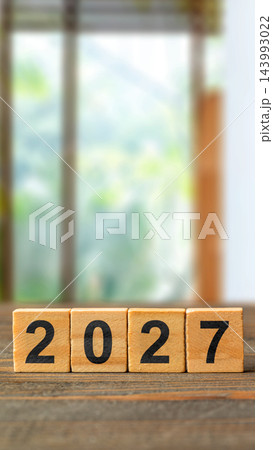 Wooden blocks spelling out the year 2027 placed on a rustic wooden table against a softly blurred, bright indoor background. Happy New Year 2027 143993022