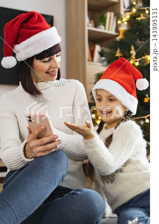 A mother and daughter wearing Santa hats share a moment looking at a smartphone together near a decorated Christmas tree A mother and daughter wearing Santa hats share a moment looking at a smartphone together near a decorated Christmas tree 143995131