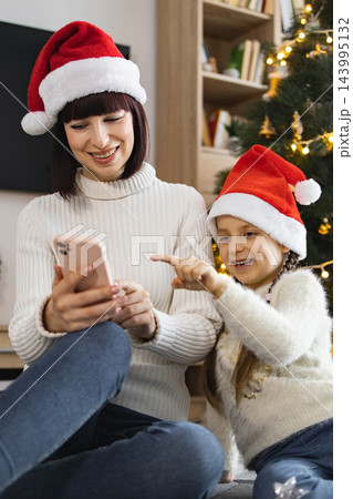 A mother and her young daughter wearing Santa hats look at a smartphone together near a decorated Christmas tree A mother and her young daughter wearing Santa hats look at a smartphone together near a decorated Christmas tree 143995132